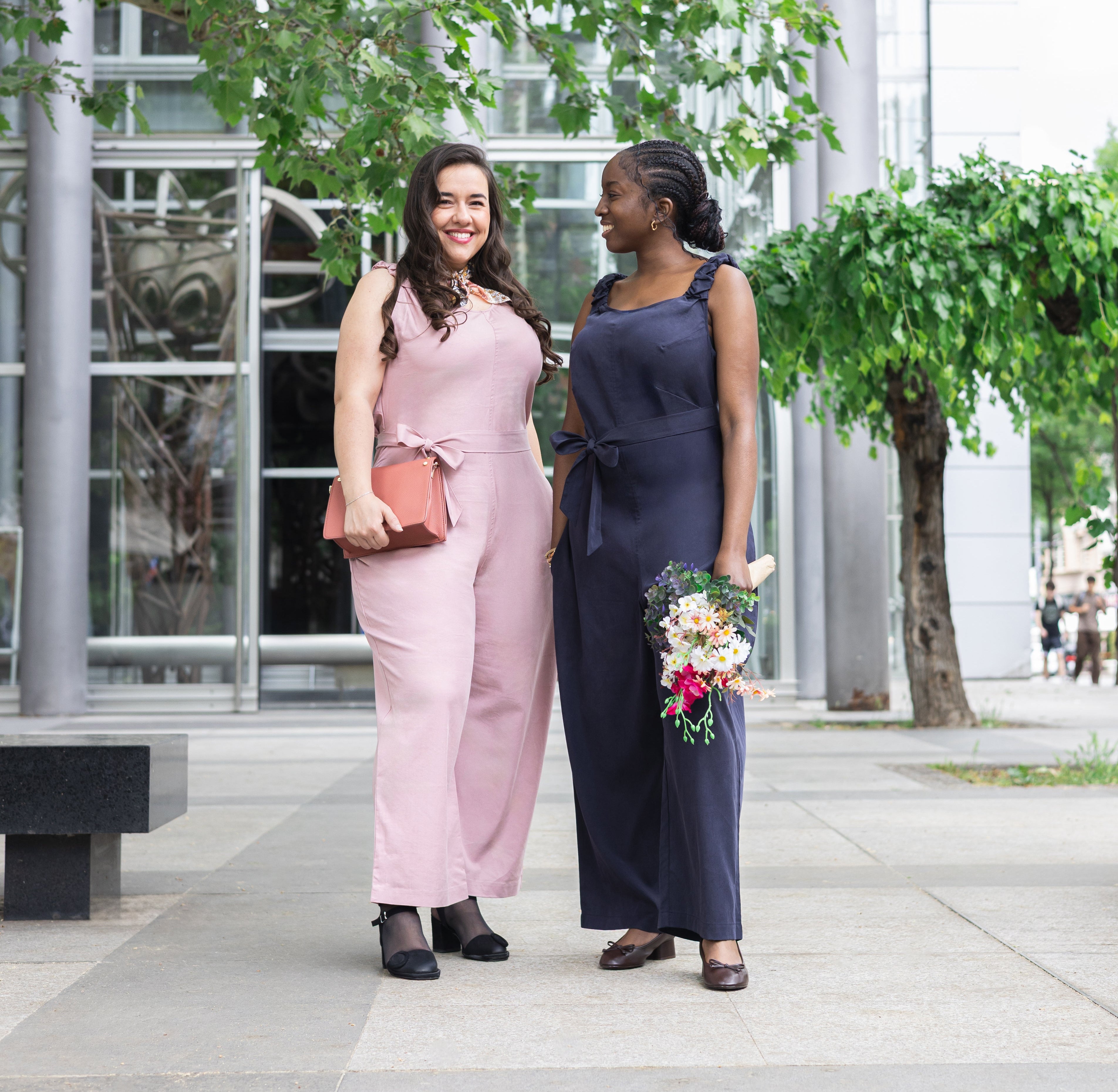 Two women in wearing the Exclusively Kristen Jumpsuit for big boobs standing outdoors with trees and a building in the background.